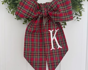 May include: A red and green plaid bow with a white embroidered letter K. The bow is tied with a loop and tails.