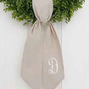 May include: A beige linen wreath bow with a white embroidered letter "D" in a script font.