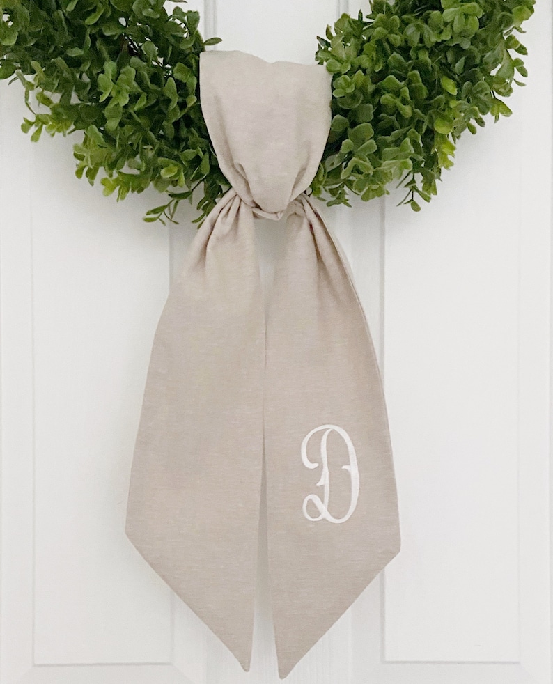 May include: A beige linen wreath bow with a white embroidered letter "D" in a script font.