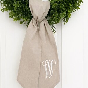 May include: A beige linen wreath bow with a white embroidered monogram "W" on the front.