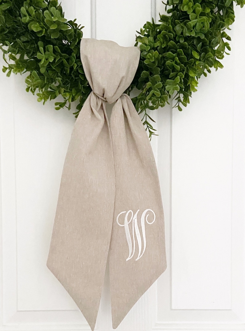 May include: A beige linen wreath bow with a white embroidered monogram "W" on the front.