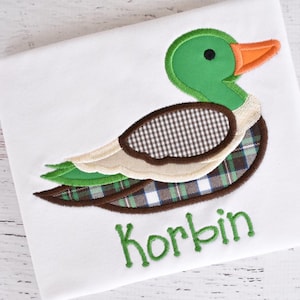 以下が含まれることがあります： 白地に緑色のアヒルが刺繍されたTシャツ。アヒルは茶色と白の格子模様のアクセントが付いています。アヒルの下に緑色の文字で「Korbin」と刺繍されています。