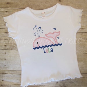 Op de afbeelding: Wit T-shirt met korte mouwen met een roze walvis afbeelding en de naam "Lola" geborduurd in blauwe, groene en roze letters.