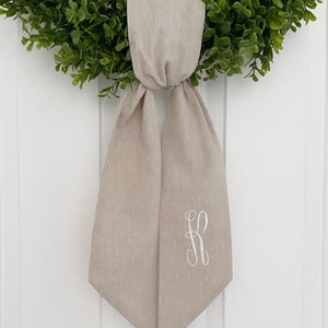 May include: A beige linen sash with a white embroidered letter "R" on the right side. The sash is tied in a bow and hangs from a green wreath.