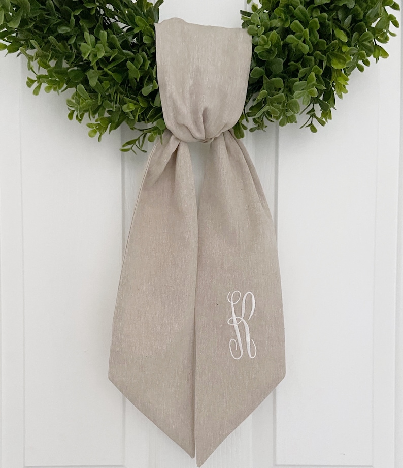 May include: A beige linen sash with a white embroidered letter "R" on the right side. The sash is tied in a bow and hangs from a green wreath.