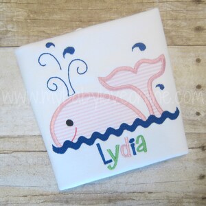 Op de afbeelding: Een wit T-shirt met een geborduurde roze walvis met blauwe golven en een blauwe waterstraal. De naam "Lydia" is in groene, roze en blauwe letters onder de walvis geborduurd.