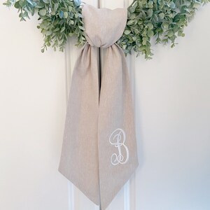 May include: A beige linen wedding sash with a white embroidered letter "B" on one side.