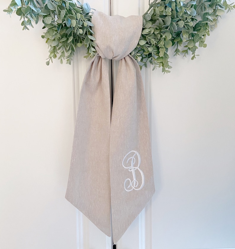 May include: A beige linen wedding sash with a white embroidered letter "B" on one side.