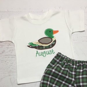以下が含まれることがあります： 緑色の刺繍されたアヒルと緑色の糸で「August」という名前が刺繍された白い半袖Tシャツ。シャツは緑と茶色の格子縞のショートパンツと組み合わされています。