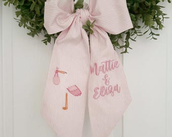 May include: Pink and white striped bow with a pink stork carrying a baby and the names Mattie & Eliza embroidered on it.