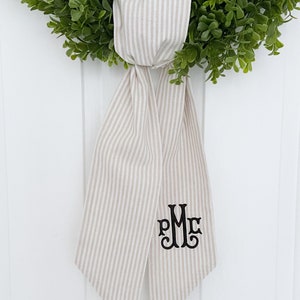 May include: A beige and white striped fabric bow with a black monogram "PMC" embroidered on it. The bow is tied around a green wreath.
