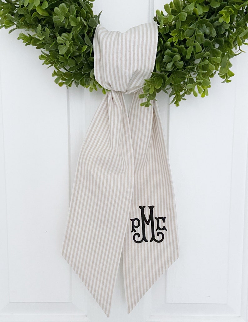 May include: A beige and white striped fabric bow with a black monogram "PMC" embroidered on it. The bow is tied around a green wreath.