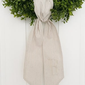 May include: A beige and white striped fabric ribbon with a large embroidered letter "B" in the center. The ribbon is tied around a green wreath.