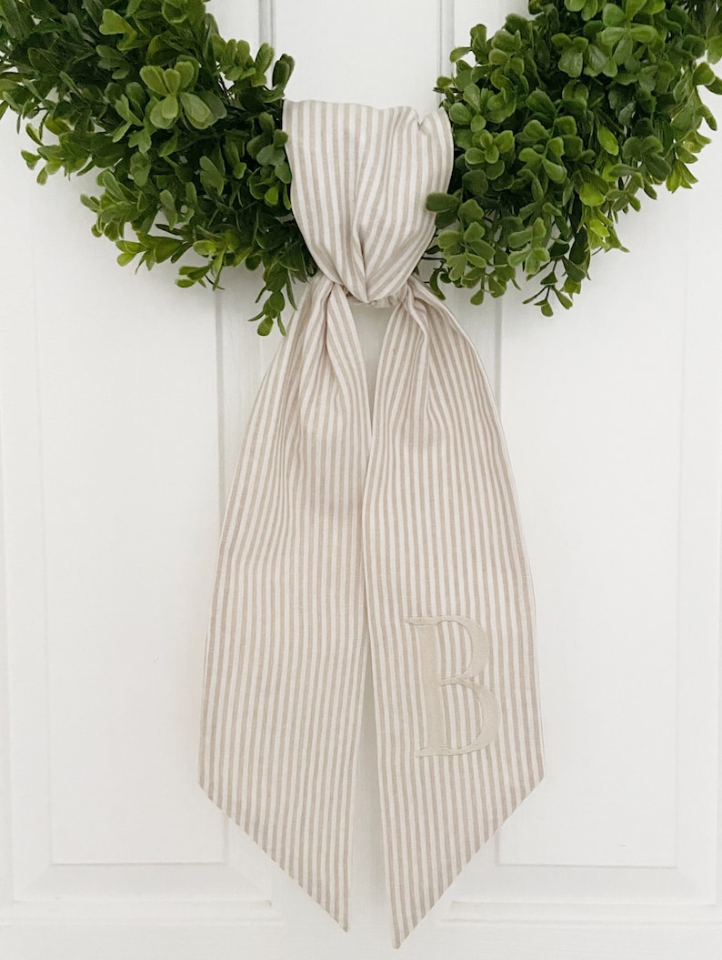 May include: A beige and white striped fabric ribbon with a large embroidered letter "B" in the center. The ribbon is tied around a green wreath.
