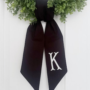 May include: A black wreath bow with a white embroidered "K" is tied to a green leafy wreath. The bow hangs down, creating a decorative accent. The wreath is displayed on a white door.