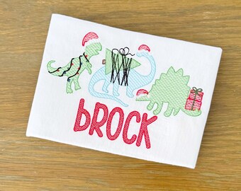 May include: White rectangular fabric with embroidered Christmas dinosaurs. One dinosaur is decorated with lights, another carries a gift, and the third has a gift. The name "BROCK" is embroidered in red.