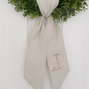 May include: A beige and white striped fabric bow with a gold embroidered monogram. The bow is tied around a green wreath.