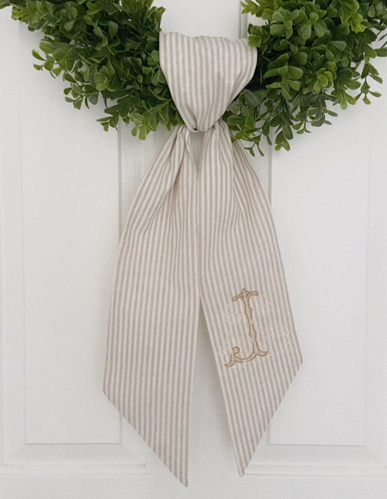 May include: A beige and white striped fabric bow with a gold embroidered monogram. The bow is tied around a green wreath.