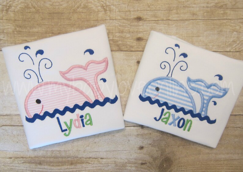 Op de afbeelding: Twee witte rompertjes met geborduurde walvisontwerpen. E&eacute;n rompertje heeft een roze walvis met de naam "Lydia" eronder geborduurd. Het andere rompertje heeft een blauwe walvis met de naam "Jaxon" eronder geborduurd.