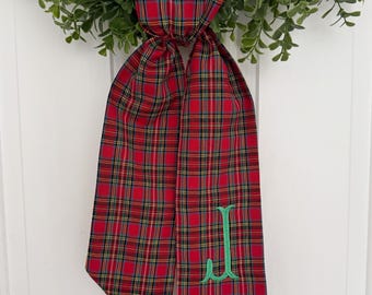 May include: Red and green plaid ribbon with a green embroidered letter "J" on it. The ribbon is tied in a bow and is hanging from a wreath.