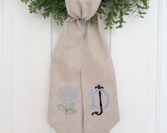 May include: A beige linen ribbon with a blue hydrangea and a black embroidered letter "D" on it. The ribbon is tied in a bow and is hanging from a green wreath.