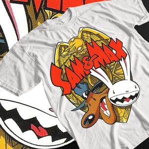 Può includere: Maglietta bianca con una stampa grafica di Sam & Max, un cane e un coniglio dei cartoni animati, davanti a un emblema di aquila dorata. Il testo "Sam & Max" è in rosso.
