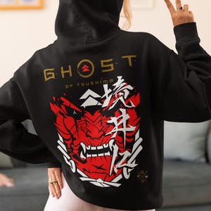 Peut inclure: Sweat-shirt à capuche noir avec un imprimé graphique blanc d'un masque de démon rouge et le texte "Ghost of Tsushima" en or.