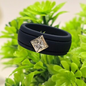 May include: A matte black silicone ring featuring a single square-cut clear gemstone set in a metallic bezel. The band has a smooth, wide profile with two horizontal grooves running along the center, creating a distinct layered appearance.