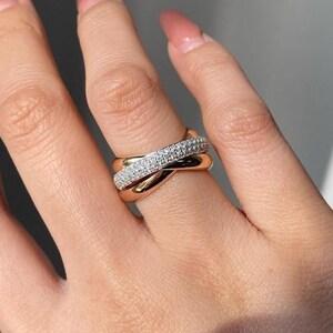 May include: A gold-toned ring with three intertwined bands. One band is encrusted with small, sparkling white stones. The other two bands are smooth and polished, creating a contrasting texture. The ring is worn on a finger.