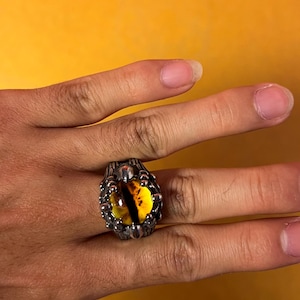 May include: A silver-colored ring with a large, oval-shaped, yellow and black eye-like stone. The ring has a detailed, textured design around the stone, and is worn on a finger. The background is a solid, bright yellow.