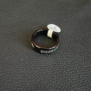 May include: Black ring with the text "HARLEY-DAVIDSON" in white lettering. The ring is size 9. The ring is on a black textured surface.