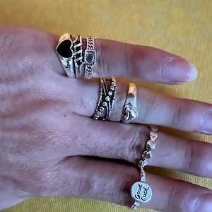 Op de afbeelding: Een verzameling zilveren ringen met diverse ontwerpen, waaronder een zwart hart, een kettingpatroon en een band met kleine steentjes. Een ring met een hartvorm en een armband met hartbedels en een "Good Luck" bedel zijn ook zichtbaar.