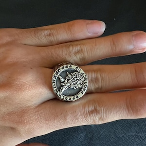 May include: A silver ring with a detailed design featuring a central emblem and surrounding text. The ring is worn on a finger, showcasing its intricate craftsmanship and bold style. The ring's design includes the words "MICHAEL PROTECT US".
