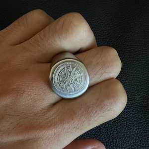 May include: A silver ring featuring a detailed compass rose design. The ring is round with a flat top, and the compass rose is intricately engraved. The ring is worn on a finger, highlighting its size and design.