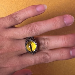 May include: A silver-colored ring featuring a large, oval-shaped yellow eye with a black slit pupil. The ring has a detailed, textured setting. The ring is shown on a hand against a yellow background.
