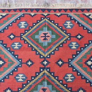 May include: A red, blue, green, and beige patterned rug with a geometric design. The rug has a fringe border.