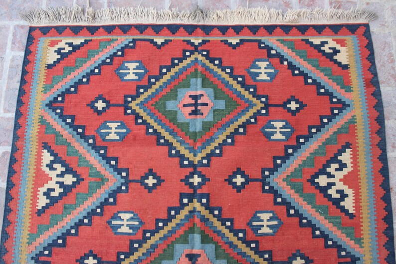 May include: A red, blue, green, and beige patterned rug with a geometric design. The rug has a fringe border.