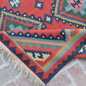 May include: A red, blue, green, and cream colored woven rug with a geometric pattern. The rug has fringe on all sides.