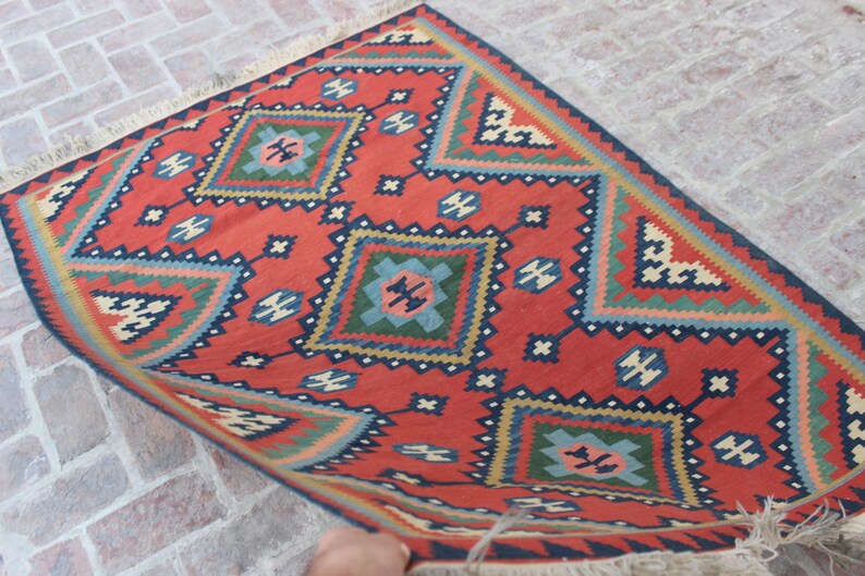 May include: A red, blue, green, and cream colored rug with a geometric pattern. The rug has a fringe border.
