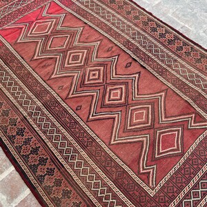 Puede incluir: Una alfombra roja y negra con un diseño geométrico. La alfombra tiene una forma de diamante central con diamantes más pequeños y zigzags que la rodean. El borde de la alfombra es un patrón repetitivo de diamantes negros y blancos.