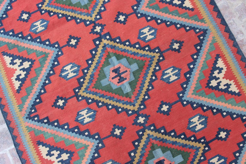 May include: A red, blue, green, and cream colored rug with a geometric pattern. The rug has a diamond shape in the center with smaller diamond shapes surrounding it.