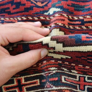May include: A close-up of a hand touching a red, blue, and white patterned rug. The rug has a traditional design with geometric shapes.