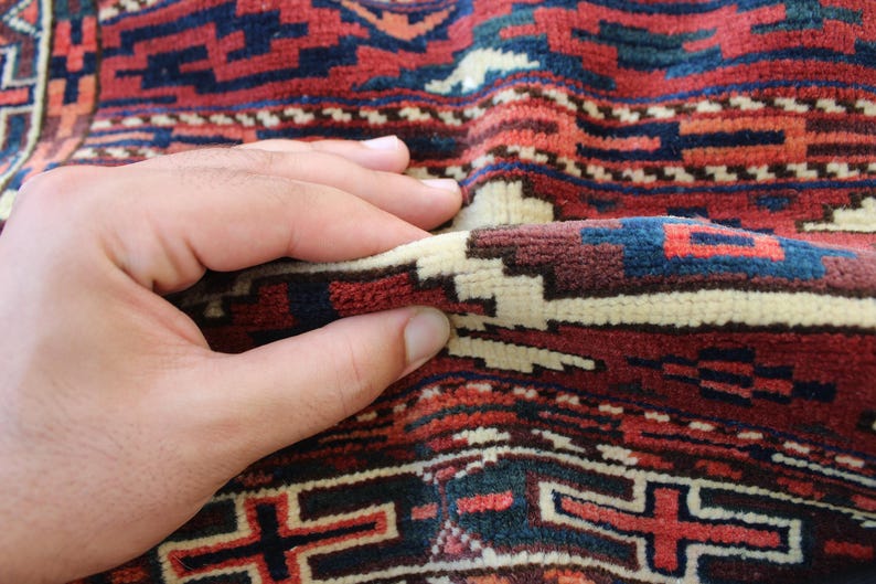 May include: A close-up of a hand touching a red, blue, and white patterned rug. The rug has a traditional design with geometric shapes.