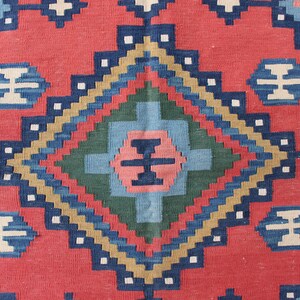 May include: A close-up of a red, blue, green, and beige patterned rug with a diamond-shaped design in the center.