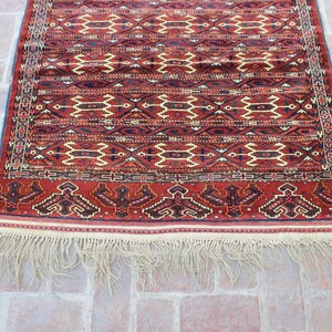 May include: A red, white, and blue patterned rug with a fringe border. The rug has a traditional design with geometric shapes and a repeating pattern.