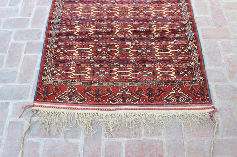 May include: A red, white, and blue patterned rug with a fringe border. The rug has a traditional design with geometric shapes and a repeating pattern.