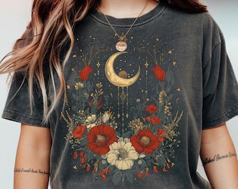 May include: Dark gray t-shirt featuring a floral and celestial design. The graphic includes a gold crescent moon, red and white flowers, and other botanical elements. A gold necklace is worn.