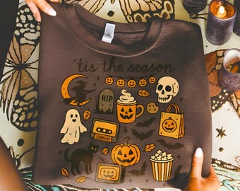 May include: A dark brown sweatshirt featuring the text "'tis the season" and Halloween illustrations. The design includes a witch on a crescent moon, a ghost, a skull, pumpkins, bats, and other festive elements.