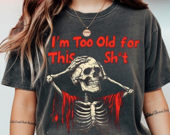 May include: Dark gray t-shirt with the text "I'm Too Old for This Sh*t" in red. The shirt features a skeleton graphic with its hands on its head and a red shawl. The shirt is a vintage style.