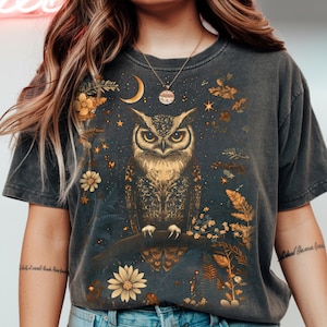 Puede incluir: Camiseta gris oscuro con una ilustración detallada de un búho posado en una rama. El diseño incluye una luna creciente, estrellas y elementos florales en tonos dorados y amarillos. La camiseta tiene un aspecto vintage.
